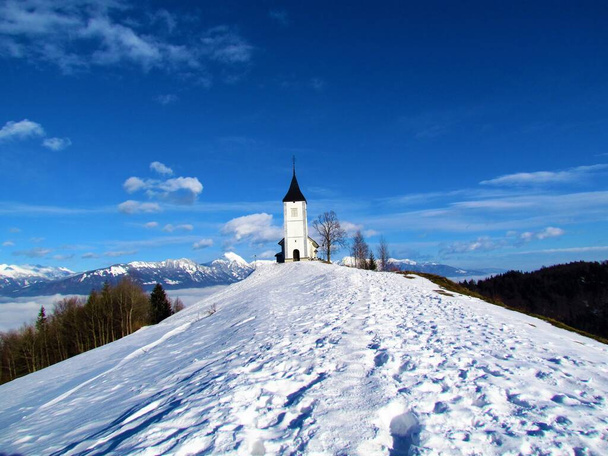 Iglesia de San Primoz y Felician en Jamnik en Gorenjska, Eslovenia en invierno con el pico de montaña nevado de Storzic - Foto, Imagen