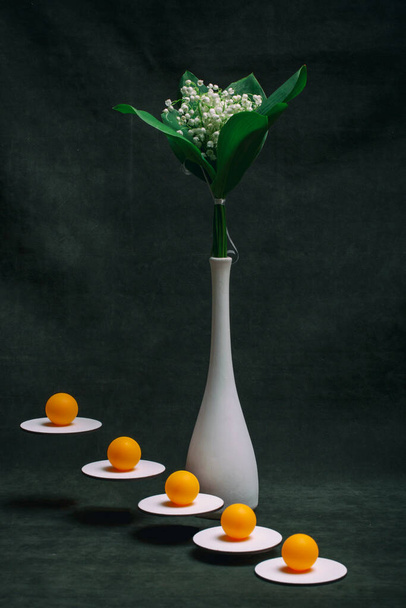 Csendélet narancssárga golyókkal a fehér állványon, és egy csokor liliom a völgyben egy fehér vázában. - Fotó, kép