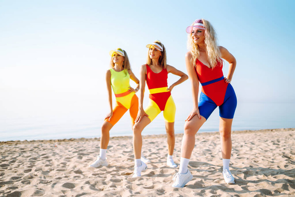 Beautiful women in swimsuits are dancing on the beach. Three woman are training in the morning. Fitness, training, aerobics and people concept. Active sport. - Фото, зображення