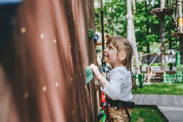 Little girl preschooler wearing safety harness training on rock climbing wall in adventure rope park. Summer camp activity for kids. - Foto, Bild