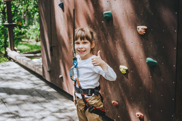 Little girl preschooler wearing safety harness giving a thumb up gesture having fun time on rock climbing wall in adventure rope park. Summer camp activity for kids. - Photo, image