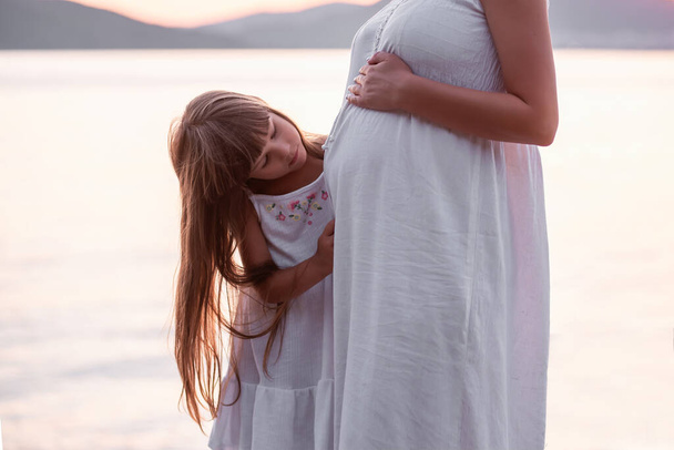 Close-up portrait of a little girl hugging pregnant mother. A daughter in a white sundress strokes a belly to a middle-aged woman by the sea at sunset. Family travel. The concept of happy motherhood - Фото, изображение