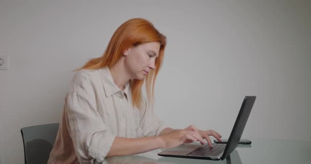 Busy woman freelancer works on laptop distracting on phone call from husband. Redhead lady takes time for quick conversation and continues performing task - Séquence, vidéo
