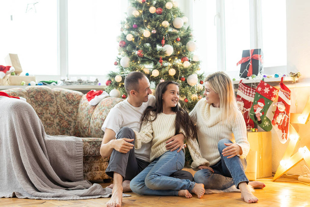 Рождественская семья Счастье. Портрет отца, мамы и дочери, сидящих дома возле елки. - Фото, изображение