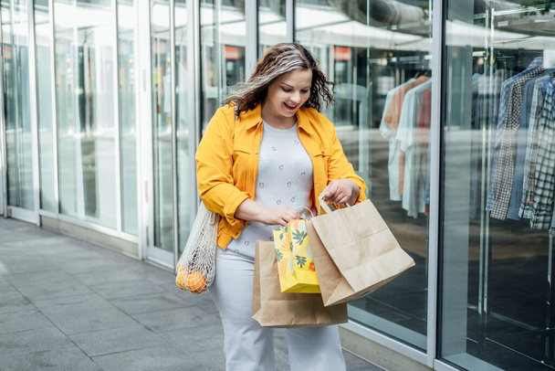 Glücklich selbstbewusst lächelnd plus Größe kurvige junge Frau mit Einkaufstaschen und trendiger Netztasche, die auf der Stadtstraße in der Nähe der Schaufenster von Einkaufszentren spaziert - Foto, Bild