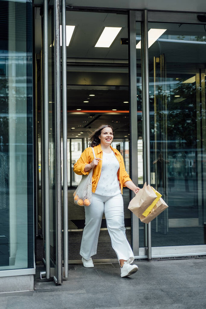ショッピングバッグや流行のネットバッグを持つ幸せな自信の笑顔プラスサイズ曲線的な若い女性がショップモールの窓の近くの街の通りを歩く - 写真・画像