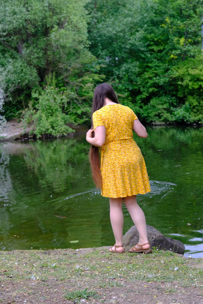 happy young woman with long hair in a yellow dress stands on the banks of a river, a lake and looks into the water, rear view, the concept of a happy female life, an active lifestyle, healthy hair - Foto, imagen