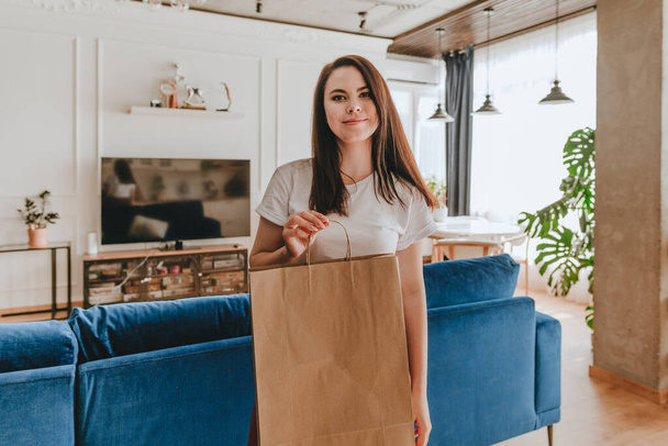 Beautiful smiling young woman holding blank craft paper bag packet package staying in the living room. Service delivery concept. - Foto, Imagem