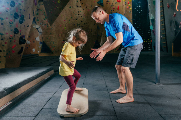 Father supporting his daughter while she training on fitness balance board in a gym. Little girl using exercise accessory rocker board. Family sport activities. Rock climbing wall on background. - Fotografie, Obrázek
