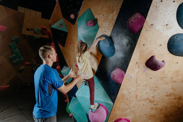 Father and his daughter rock climbers training in a modern indoor rock climbing gym. Male instructor helps little girl preschooler on a private coach class. Family sport activities. Selective focus. - Foto, Bild