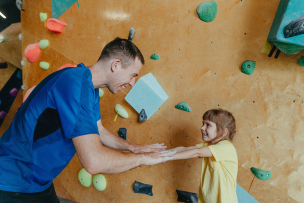 Father and his daughter rock climbers training in a modern indoor rock climbing gym. Male instructor giving high five to little girl preschooler on a private coach class. Family sport activities. Selective focus. - Foto, Bild