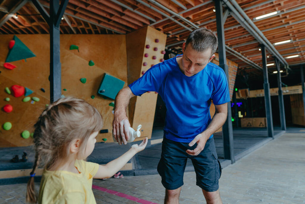 Father and his daughter training in a modern indoor rock climbing gym. Male instructor giving little girl preschooler liquid chalk used to improve grip. Family sport activities. Selective focus. - Foto, afbeelding