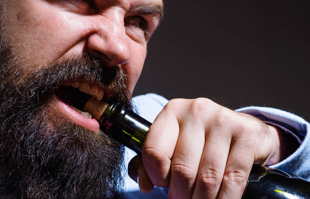 Een man met baard die een fles champagne of wijn probeert te openen. De man trekt kurk met tanden eruit. Sluitingsdatum - Foto, afbeelding