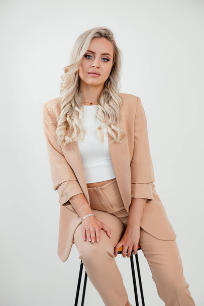 Blonde cote long hair business Woman wearing stylish formal wear beige suit on white background, sitting on chair, studio shot, look book  - Photo, Image