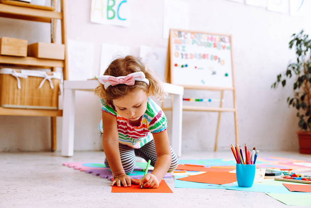 Klein meisje met mooie bezel op het hoofd zitten op de vloer en tekening met gekleurde potloden op vel papier in de kleuterschool. Interessante les in speelkamer voor kleuters die creatie ontwikkelen. - Foto, afbeelding