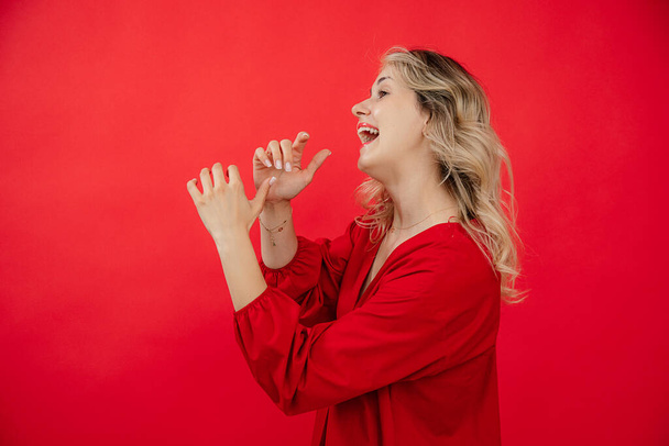 Portrait side view of cheerful young woman in red outfit playing on invisible instrument isolated on red background. Sing and speak into mouthpiece, loudspeaker. Fashion and beauty industry. - Photo, Image