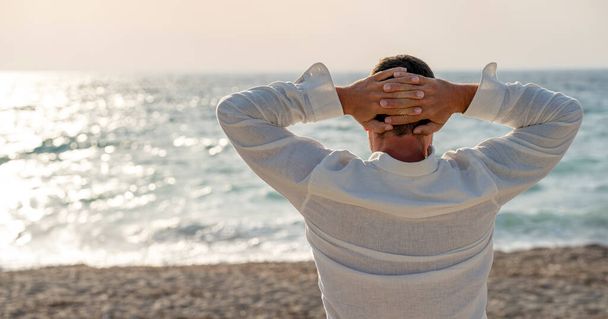 Vacaciones en la playa. Vista trasera de un joven relajado con camisa blanca disfrutando de la vista al mar - Foto, Imagen