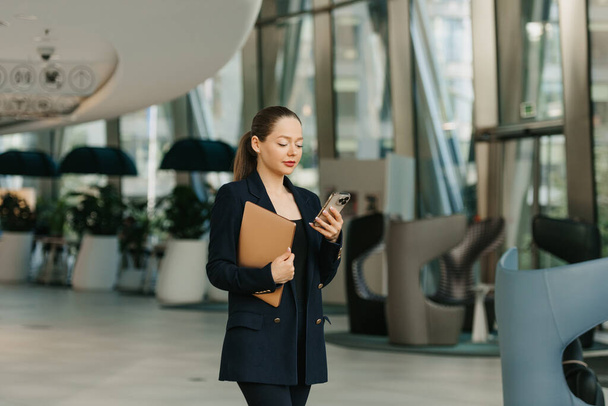 A female realtor is holding a laptop and enjoying her cell phone. A pretty woman is standing in the modern lobby. A businesswoman is leaving her office in a contemporary interior. - Foto, afbeelding
