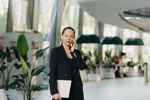 A female realtor who is holding a laptop and having a conversation on a smartphone. A woman is standing in the modern lobby. A business woman is leaving her office in a contemporary interior. - Photo, Image