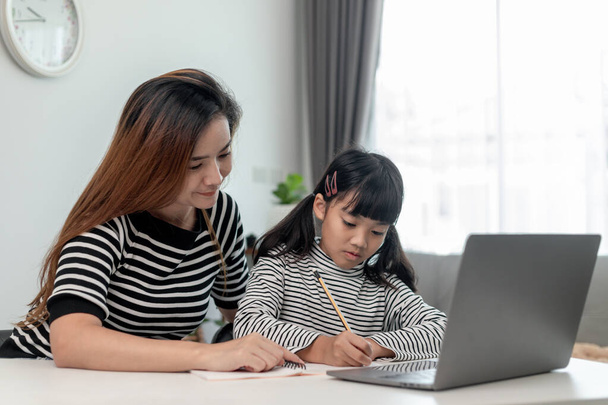 Asiatique petite fille enfant apprendre classe en ligne à la maison avec la mère. L'ordinateur portatif d'utilisation d'enfant préscolaire font des devoirs, l'école à domicile de l'enseignant d'école par l'Internet à distance numérique avec le soutien de maman. - Photo, image