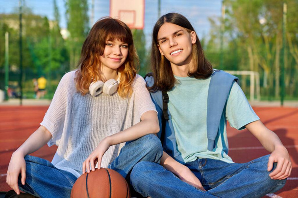 Portrait of couple friends teenagers guy and girl looking at camera, sitting on school basketball court. Teenage students with backpacks, ball in sunset. Active healthy sport lifestyle, youth concept - Foto, immagini