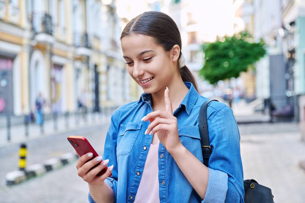 Beautiful teenage girl talking using smartphone, urban background. Attractive positive emotional young female looking at phone on city street. Technology leisure communication lifestyle youth concept - Photo, image