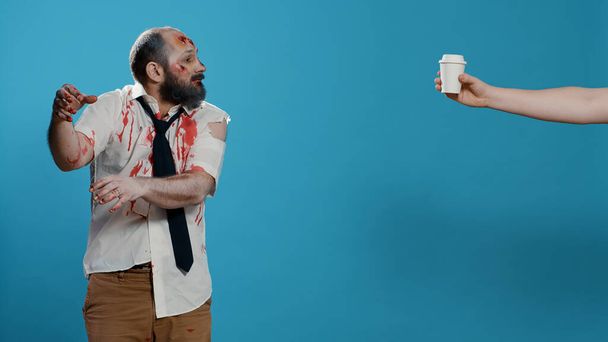 Beyinsiz beyin yiyen zombi mavi arka planda bir fincan kahve almaya çalışıyor. Tehlikeli kıyamet beyin ölü yürüyen ceset homurdanarak kahve fincanını kapmaya çalışıyor. Stüdyo çekimi - Fotoğraf, Görsel