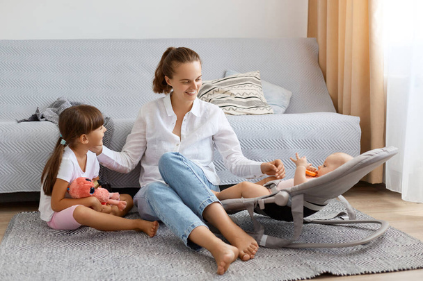 Indoor shot of satisfied attractive woman with ponytail wearing white shirt and jeans sitting on floor with her children, playing with infant baby in rocking chair together with elder female kid. - Zdjęcie, obraz