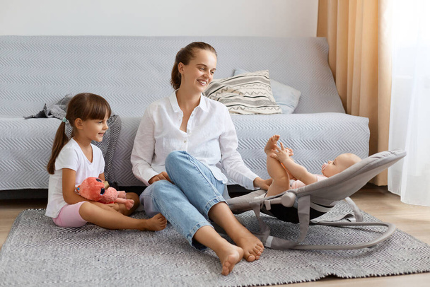 Horizontal shot of adorable delighted woman wearing white shirt and jeans sitting on floor with her kids, spending time with with toddler kid in rocking chair together with elder child. - Photo, image