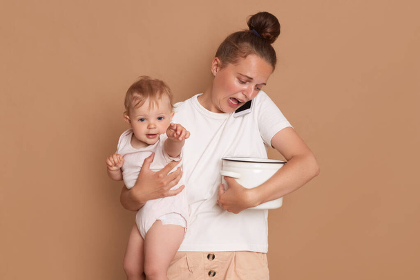 Indoor shot of exhausted shocked woman wearing white casual style shirt holding in arms toddler daughter, looks at the spoiled food in the pot while talking on smartphone isolated on brown background. - Фото, зображення