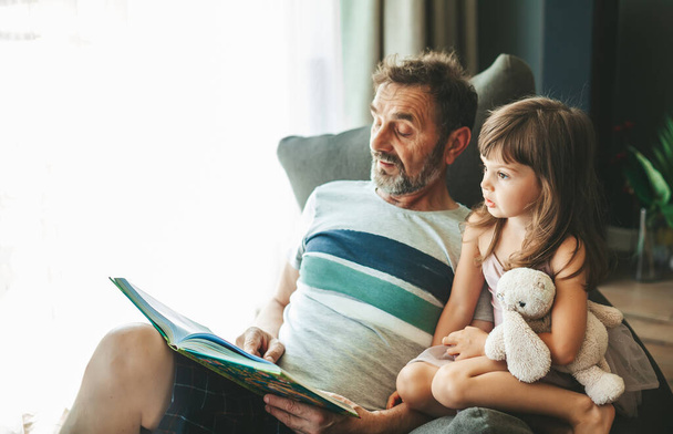 Isoisä lukee kirjaa tyttärentyttärelleen, istuu papupussituolilla - Valokuva, kuva