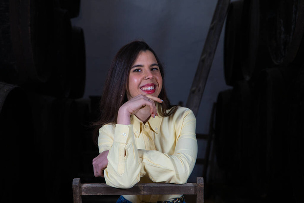 Young beautiful businesswoman sitting on a chair in her wine cellar. The woman is making different expressions and gestures looking at the camera. Enterprising woman concept - Photo, Image