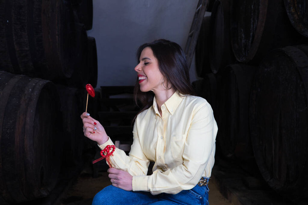 Young beautiful businesswoman sitting on a chair in her wine cellar. The woman is making different expressions and gestures while holding hearts and looking at the camera. Enterprising woman concept. - Photo, Image