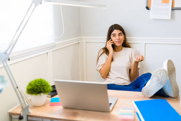 Busy casual young woman at home talking on the phone with a client while relaxing and working at her sunny office  - 写真・画像