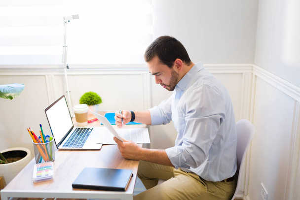 Akıllı bir genç adam kontratı kontrol ediyor ve ofisinde çalışıyor. Masasında oturan beyaz bir adamın yan görüntüsü.  - Fotoğraf, Görsel