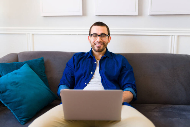 Portret van een gelukkige man met een bril die lacht en typt op de laptop terwijl hij freelance werk doet in de bank thuis  - Foto, afbeelding