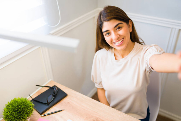 Perspectiva personal de una mujer atractiva sonriendo y tomando una selfie en su oficina mientras trabaja desde casa - Foto, imagen