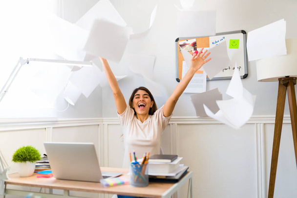 Excited young woman throwing papers and celebrating finishing her work while sitting at her desk at home - Foto, immagini
