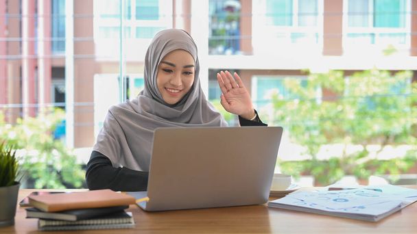 Jovem mulher de negócios muçulmana no hijab tendo videochamada on-line no laptop enquanto está sentado no local de trabalho moderno. - Foto, Imagem