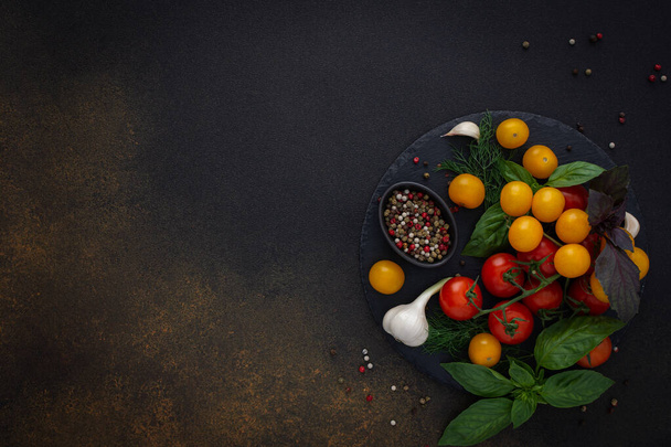 Ruokatausta, tuoreet kypsät punaiset ja keltaiset tomaatit, mausteet ja basilikan lehdet, valkosipuli ja vihreät sipulit tummalla laudalla, terveellisen ruoan konsepti, kopiotila, ylhäältä katsottuna - Valokuva, kuva