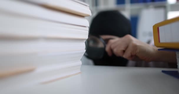 La persona in passamontagna con lente d'ingrandimento guarda documenti in ufficio. Concetto di spionaggio industriale e furto di proprietà intellettuale - Filmati, video