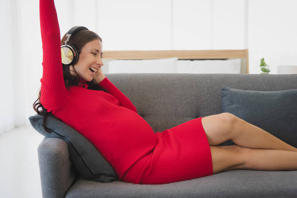 Kırmızı kazaklı hamile kadın kanepeye uzanıp modern oturma odasında dinlenmek için müzik dinliyor.. - Fotoğraf, Görsel