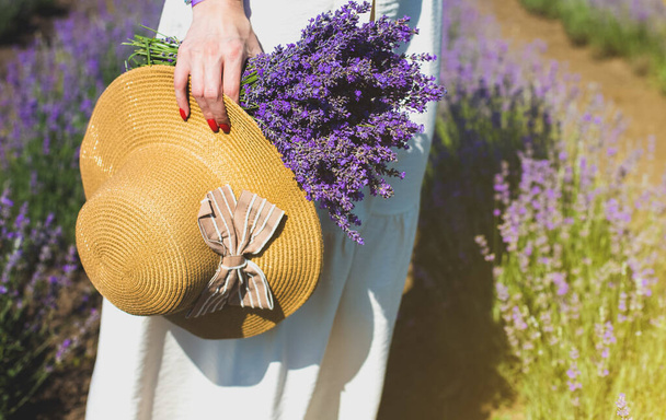Chica en vestido de lino blanco con ramo de lavanda y sombrero de paja caminando en hermosas flores de lavanda púrpura en el campo de verano. Concepto cálido e inspirador. Wanderlust y viajar. - Foto, Imagen