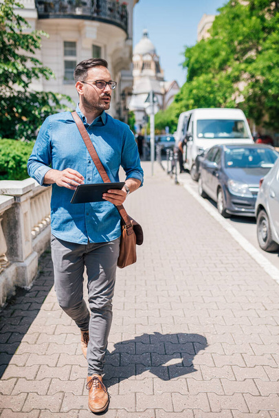 Homme d'affaires confiant avec tablette numérique détournant les yeux. Homme banlieusard professionnel marche en véhicules sur le trottoir. Il porte des formals en ville pendant la journée ensoleillée. - Photo, image