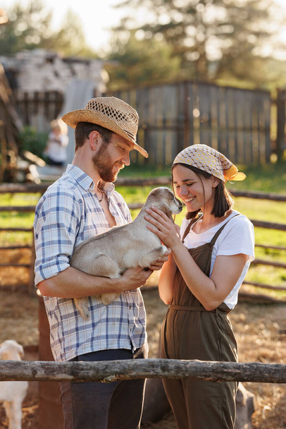 Portrait of young Caucasian happy farmer couple holding a little goat in their arms. Family business at ecological farm. Healthy food.Industrial production of goat milk dairy products. Cattle farming - Photo, Image