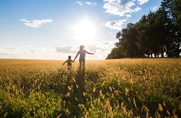Kind und Frau, Mutter und Sohn, laufen über eine gelbe Wiese der Sonne entgegen. Glückliche Kindheit, viel Spaß beim Spazierengehen. Vorwärts in eine strahlende Zukunft. Rückseite. Hintergrundbeleuchtung und weicher Fokus - Foto, Bild