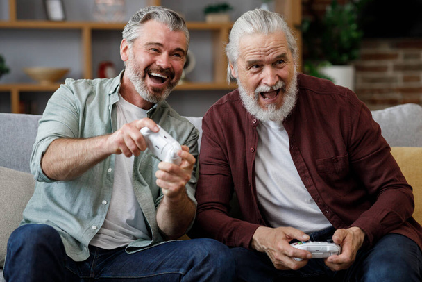 Adult son and senior father having fun, enjoy spending time together. Two generations family playing video games using gamepads at home. Fathers Day. Active modern elderly people - Foto, Imagen