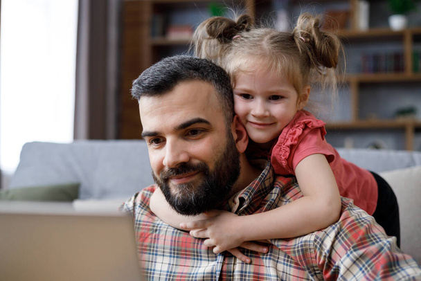 Happy family portrait. Young handsome man works from home at laptop, focus attention on laptop screen while his little daughter hugging her beloved dad around neck. Family love and family bond concept - 写真・画像