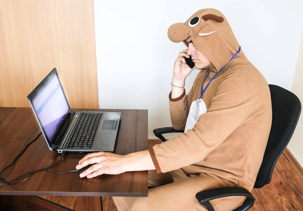 Büroangestellte im Cosplay-Kostüm einer Kuh. Kerl im lustigen Schlafanzug im Schlafanzug telefoniert. Parodie auf einen verzweifelten Manager am Laptop. Burnout am Arbeitsplatz. - Foto, Bild