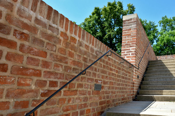 Die rote Treppe führt unter das Gelände eines Rasenplatzes mit Parkwegen hinab. Die Wände bestehen aus bearbeiteten Ziegelsteinen und Betonstufen. Hintergrundbeleuchtung Treppen mit Halogenen an den Seiten - Foto, Bild